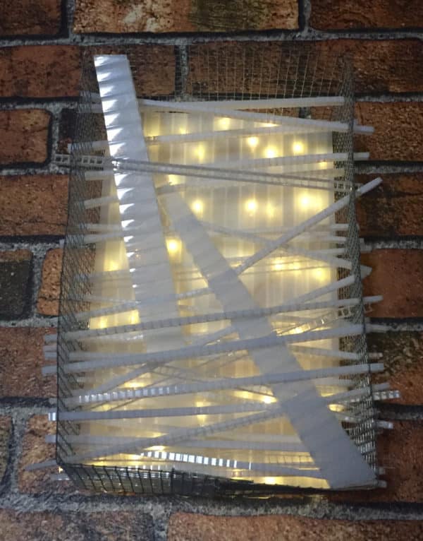 Close-up of Box #1 original Lumencrafter wall sconce light sculpture lit up, mounted on brick wall.  Rectangular white battery-powered LED light sculpture, soft white fairy lights shine behind a diffusion plate framed by a sturdy steel grid with several thin twinwall strips poking through, up and down, back and forth.