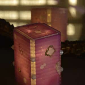 View from top, close-up of free-standing LED table light by Lumencrafter, light pink with golden splotches on tabletop by mirror.  Light yet sturdy, 6 inches high by four inch square base. Hand-painted plates of polycarbonate enclose dew drop LEDs wrapped around an inner polycarbonate core.  Indoor/outdoor. Suitable for evening al fresco dining or bedside mood lighting. Cordless; 3 AA batteries included.