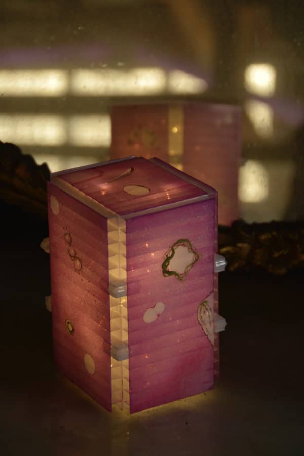 View from top, close-up of free-standing LED table light by Lumencrafter, light pink with golden splotches on tabletop by mirror.  Light yet sturdy, 6 inches high by four inch square base. Hand-painted plates of polycarbonate enclose dew drop LEDs wrapped around an inner polycarbonate core.  Indoor/outdoor. Suitable for evening al fresco dining or bedside mood lighting. Cordless; 3 AA batteries included.