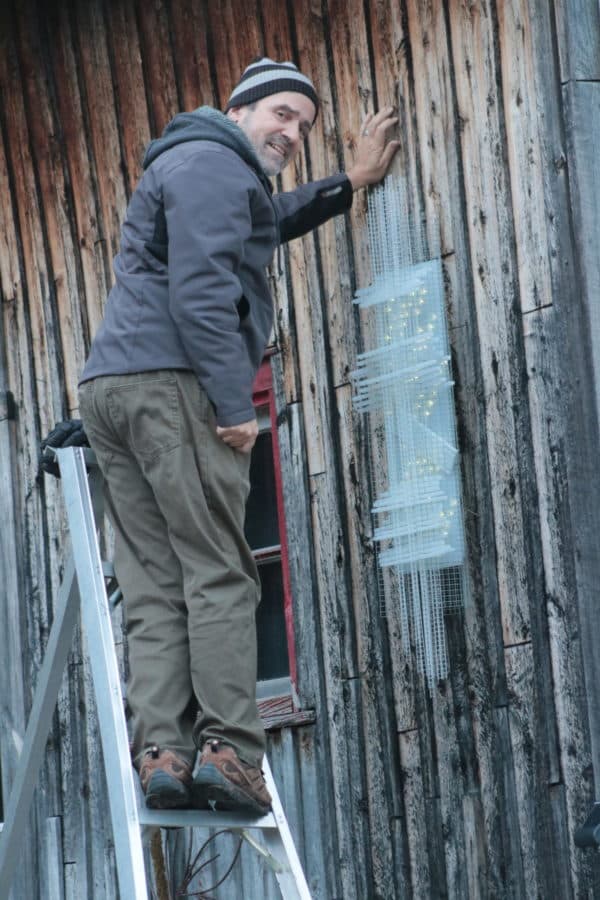 Lumencrafter Sculptor Reid Dennison attaching Solar Thing, unique solar-powered hand-made pale green four-foot long LED light sculpture to the barn at Flesherton's Local Colour's 2020 Art Garden.
