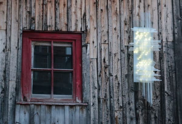 Solar Thing, unique solar-powered hand-made pale green four-foot long LED light sculpture by Lumencrafter mounted beside a red-framed window on the barn at Flesherton's Local Colour's 2020 Art Garden.