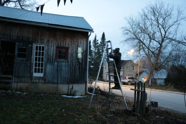 Lumencrafter Sculptor Reid Dennison checks the alignment of Solar Thing, a unique solar-powered hand-made pale green four-foot long LED light sculpture mounted on the barn at Flesherton's Local Colour's 2020 Art Garden.
