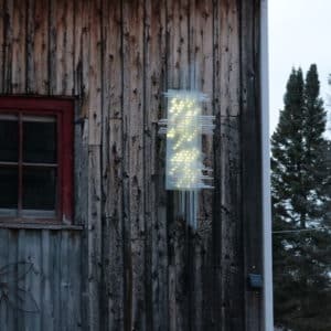 Solar Thing, unique solar-powered hand-made pale green four-foot long LED light sculpture by Lumencrafter mounted on the barn at Flesherton's Local Colour's 2020 Art Garden.