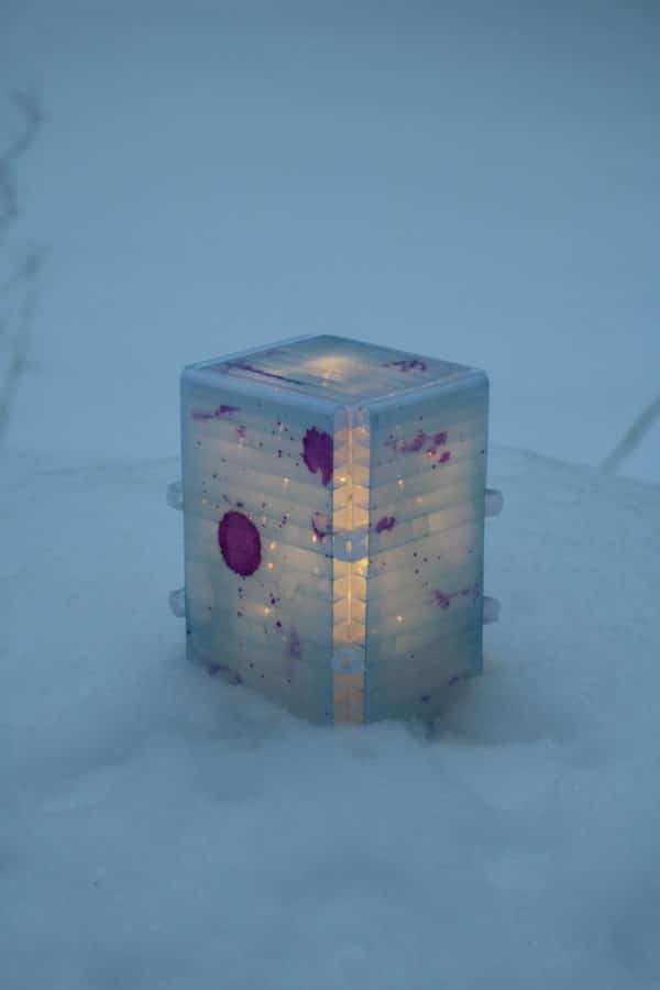 Light yet sturdy free-standing LED table light by Lumencrafter, light blue with deep pink splotches, close-up in snow, 6 inches high by four inch square base. Hand-painted plates of polycarbonate enclose dew drop LEDs wrapped around an inner polycarbonate core.  Indoor/outdoor. Suitable for evening al fresco dining or bedside mood lighting. Cordless; 3 AA batteries included.