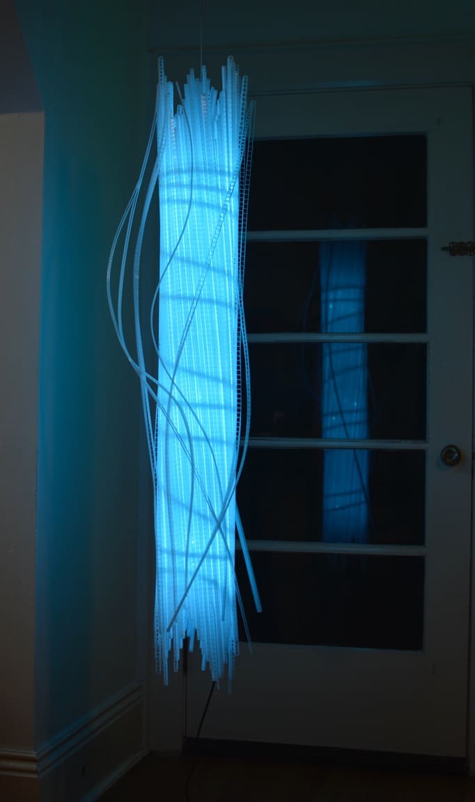 City of Light II, sky-blue 4-foot-long cylinder pendant, original LED light sculpture by Lumencrafter, reflected in glass door. strips of polycarbonate twinwall swirl around acrylic tube.