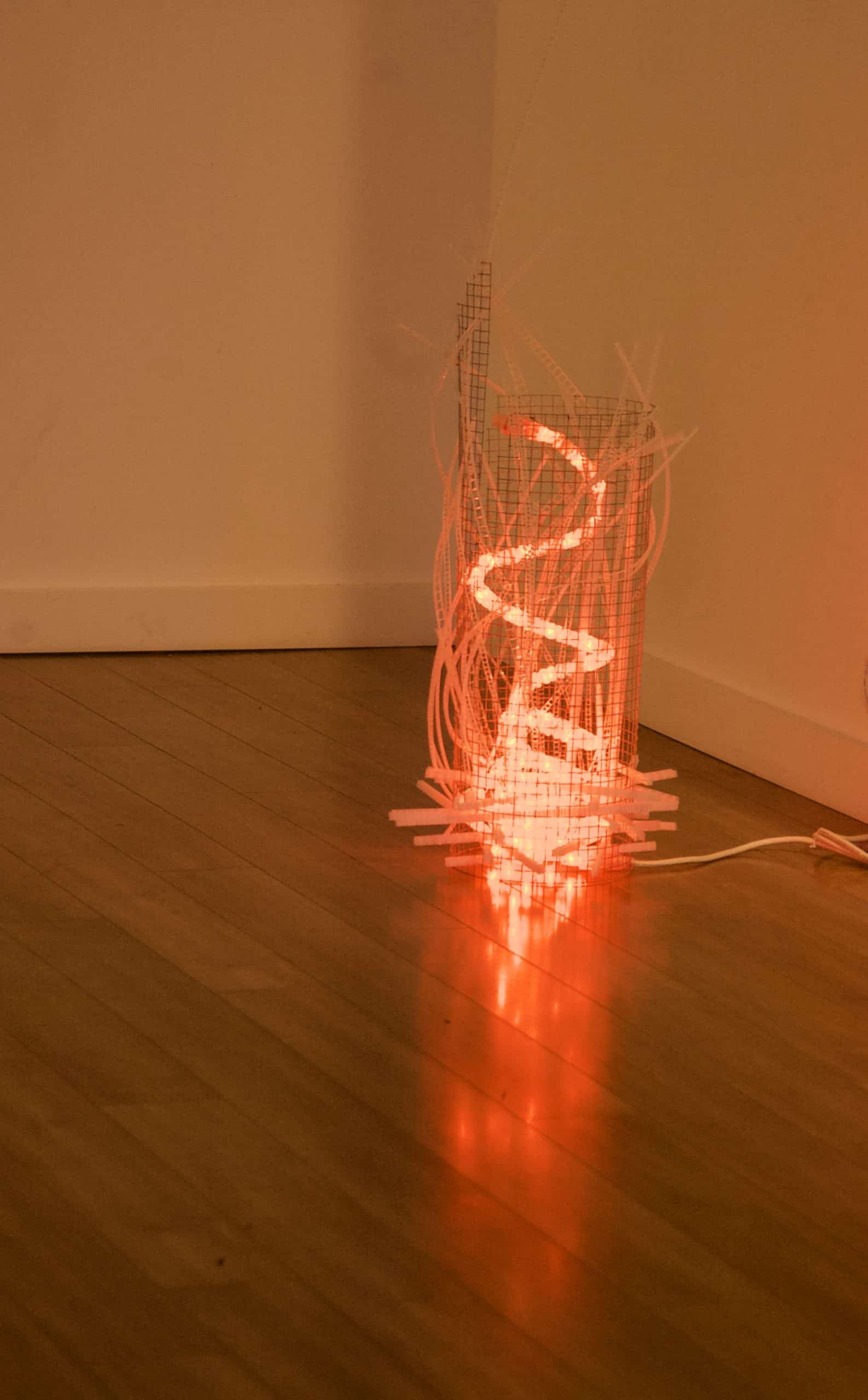 Campfire, freestanding red-orange original handmade Lumencrafter LED light sculpture, a swirling fiery rope light contained in a wire mesh cylinder shot through with strips of laddered polycarbonate twinwall on floor at Arcadia Art Gallery, Toronto, 2014.