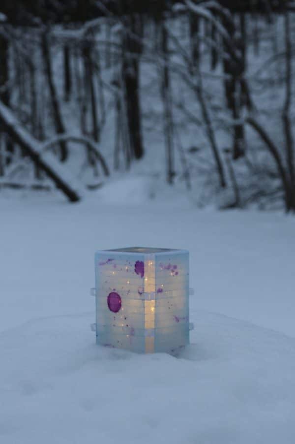 Light yet sturdy free-standing LED table light by Lumencrafter, light blue with deep pink splotches, resting on a snow-covered rock with woods in background.  6 inches high by four inch square base. Hand-painted plates of polycarbonate enclose dew drop LEDs wrapped around an inner polycarbonate core.  Indoor/outdoor. Suitable for evening al fresco dining or bedside mood lighting. Cordless; 3 AA batteries included.