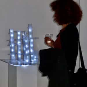 Art lover gazes at This Years' Model, a.k.a If You Lived Here, You'd be Home by Now, free-standing original handmade Lumencrafter LED light sculpture, square columns of different heights connected by swirls of laddered polycarbonate twinwall strips, glowing blue-white at Arcadia Art Gallery, Toronto, 2014.