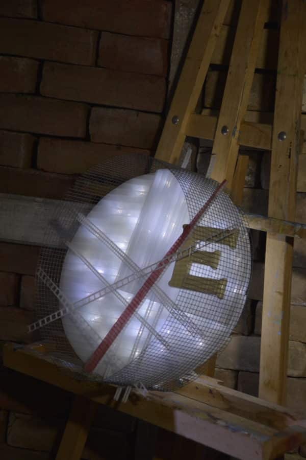 Runnin' On, original circular LED wall sconce light sculpture: A cool white LED rope light shines through a diffusion circle with large ceramic E and painted red pointer; polycarbonate strips poke through surrounding steel mesh cylinder. Sculpture rests comfortably on painter's easel.