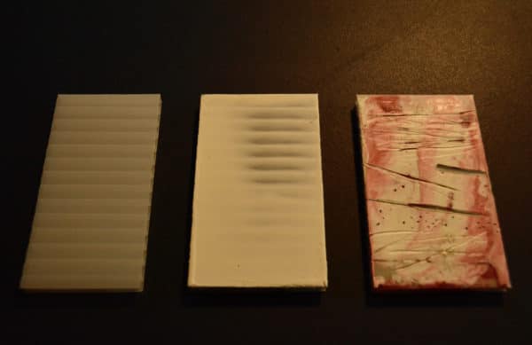 Three panels of free-standing LED table light by Lumencrafter in stages of painting: [1] pale white polycarbonate panel; [2] panel with base of white soy wax; [3] finished panel with pink and white encaustic paint, with carved-in lines and dots.  Light yet sturdy, 6 inches high by four inch square base. Hand-painted plates of polycarbonate enclose dew drop LEDs wrapped around an inner polycarbonate core.  Indoor/outdoor. Suitable for evening al fresco dining or bedside mood lighting. Cordless; 3 AA batteries included.