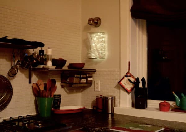 Box #1 original Lumencrafter light sculpture mounted on kitchen wall corner by stovetop.  Rectangular white battery-powered LED light sculpture, soft white fairy lights shine behind a diffusion plate framed by a sturdy steel grid with several thin twinwall strips poking through, up and down, back and forth.