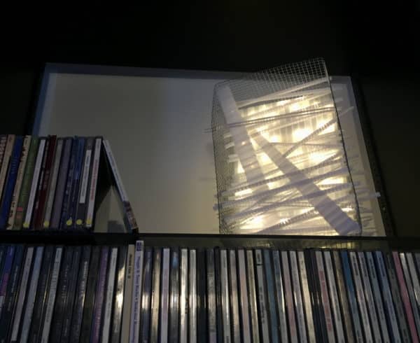 Rectangular white battery-powered LED light sculpture from Lumencrafter, soft white fairy lights shine behind a diffusion plate framed by a sturdy steel grid with several thin twinwall strips poking through, up and down, back and forth.  Free-standing sculpture set on a bookshelf by a row of CDs.