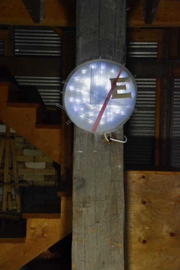 Runnin' On, original circular LED wall sconce light sculpture: A cool white LED rope light shines through a diffusion circle with large ceramic E and painted red pointer; polycarbonate strips poke through surrounding steel mesh cylinder. Sculpture mounted on large wooden post in workshop.