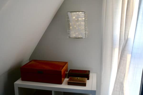 Box #1 original Lumencrafter wall sconce light sculpture mounted on bedroom wall above shelf of wooden boxes.  Rectangular white battery-powered LED light sculpture, soft white fairy lights shine behind a diffusion plate framed by a sturdy steel grid with several thin twinwall strips poking through, up and down, back and forth.