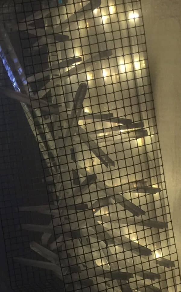 Extreme close-up, side view through steel grid, of Box #1 original Lumencrafter wall sconce light sculpture.  Rectangular white battery-powered LED light sculpture, soft white fairy lights shine behind a diffusion plate framed by a sturdy steel grid with several thin twinwall strips poking through, up and down, back and forth.