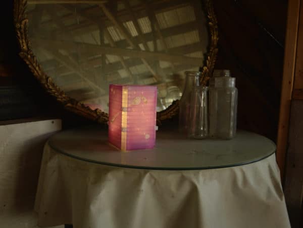 Free-standing LED table light by Lumencrafter, light pink with golden splotches on tabletop by mirror and glass bottles.  Light yet sturdy, 6 inches high by four inch square base. Hand-painted plates of polycarbonate enclose dew drop LEDs wrapped around an inner polycarbonate core.  Indoor/outdoor. Suitable for evening al fresco dining or bedside mood lighting. Cordless; 3 AA batteries included.