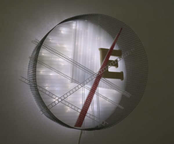 Runnin' On, original circular LED wall sconce light sculpture: A cool white LED rope light shines through a diffusion circle with large ceramic E and painted red pointer; polycarbonate strips poke through surrounding steel mesh cylinder. Sculpture mounted on white wall; front view.
