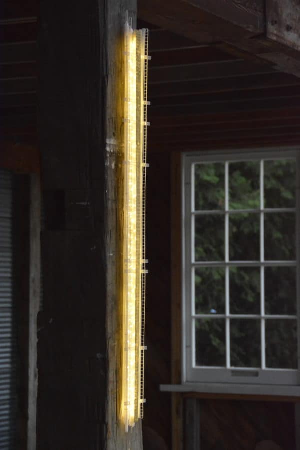 A one-inch wide strip of gold runs down the centre of a slender four-foot-long structure of polycarbonate struts and spars shot through with sparkling soft-white light.   LED light sculpture mounted on rough-hewn wooden post.