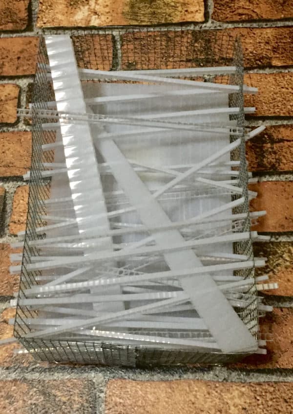 Close-up of unlit Box #1 original Lumencrafter wall sconce light sculpture mounted on brick wall.  Rectangular white battery-powered LED light sculpture, soft white fairy lights shine behind a diffusion plate framed by a sturdy steel grid with several thin twinwall strips poking through, up and down, back and forth.