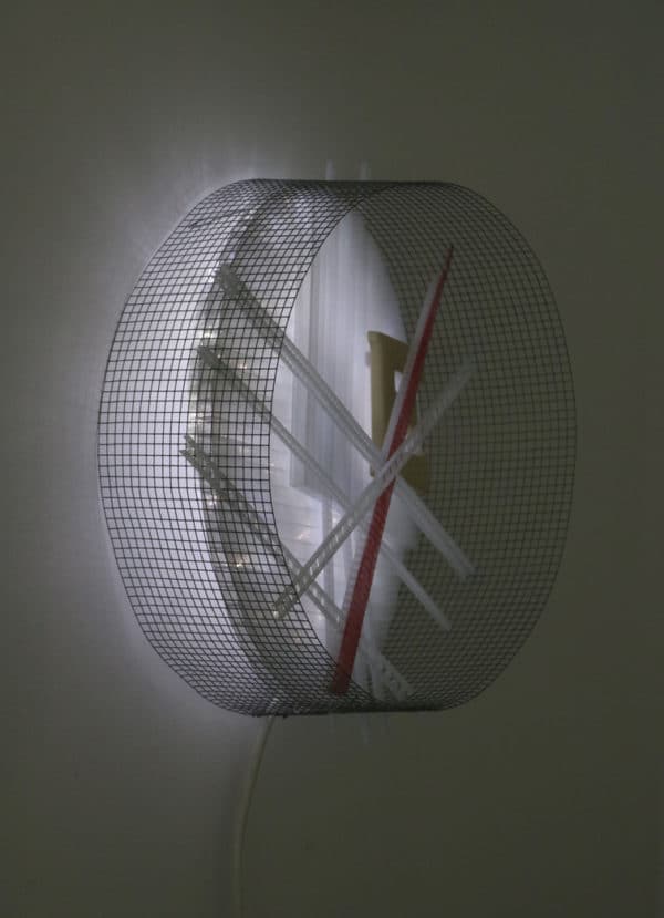 Runnin' On, original circular LED wall sconce light sculpture: A cool white LED rope light shines through a diffusion circle with large ceramic E and painted red pointer; polycarbonate strips poke through surrounding steel mesh cylinder. Sculpture mounted on white wall, side view.