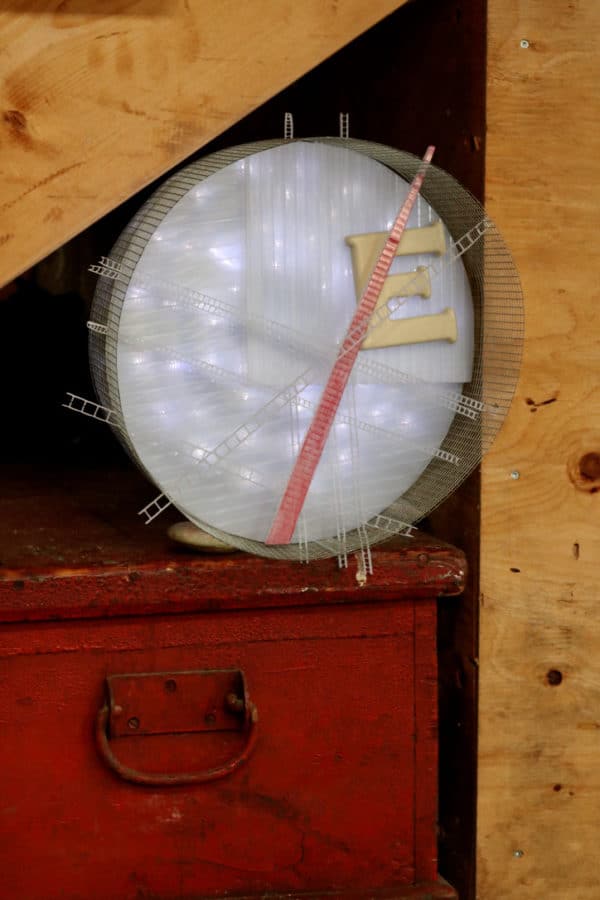 Runnin' On, original circular LED wall sconce light sculpture: A cool white LED rope light shines through a diffusion circle with large ceramic E and painted red pointer; polycarbonate strips poke through surrounding steel mesh cylinder. Sculpture rests on red wooden chest under stairs.