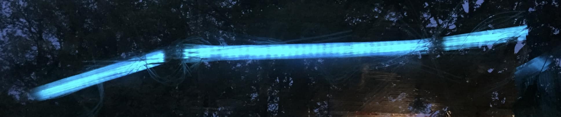 tightly cropped landscape view of A Roar, A Boar y Alice, reflected in the glass of the garage door at Sourceworks Ceramics Studio driveshed. A 22-foot-long original LED light sculpture by Lumencrafter, sky-blue LED lights run through three acrylic tubes joined with heavy gauge wire, wrapped with strips of steel mesh and polycarbonate. stunning; completely original.