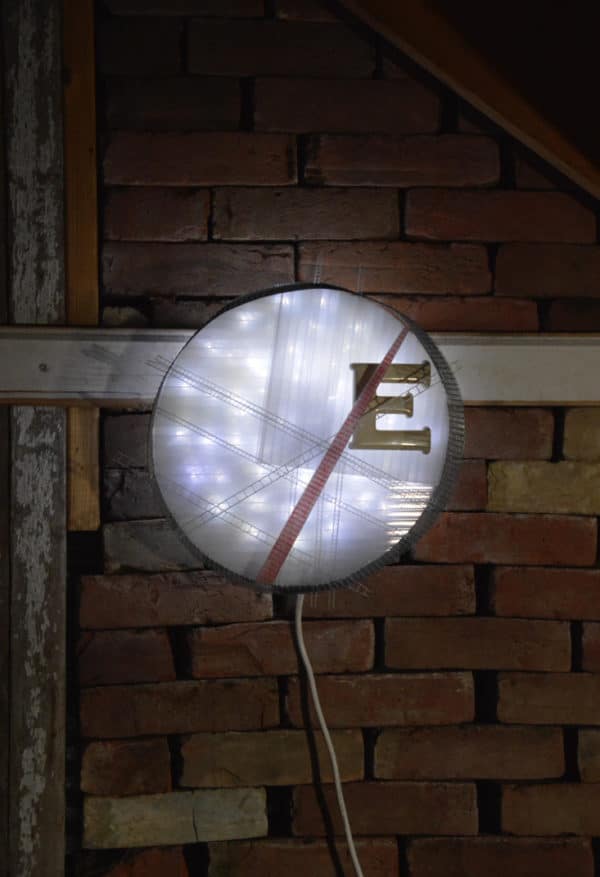 Runnin' On, original circular LED wall sconce light sculpture: A cool white LED rope light shines through a diffusion circle with large ceramic E and painted red pointer; polycarbonate strips poke through surrounding steel mesh cylinder. Sculpture mounted on brick wall.