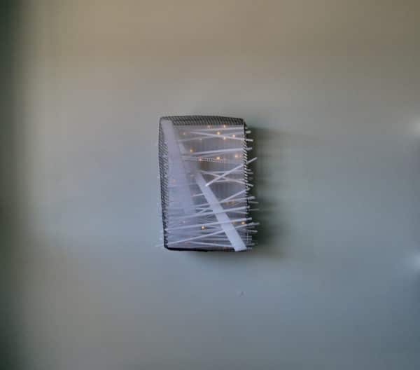 Close-up of Box #1 original Lumencrafter wall sconce light sculpture mounted on pale green wall.  Rectangular white battery-powered LED light sculpture, soft white fairy lights shine behind a diffusion plate framed by a sturdy steel grid with several thin twinwall strips poking through, up and down, back and forth.