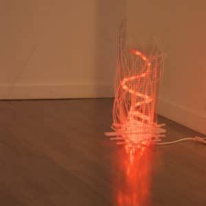 Campfire, freestanding red-orange original handmade Lumencrafter LED light sculpture, a swirling fiery rope light contained in a wire mesh cylinder shot through with strips of laddered polycarbonate twinwall on floor at Arcadia Art Gallery, Toronto, 2014.