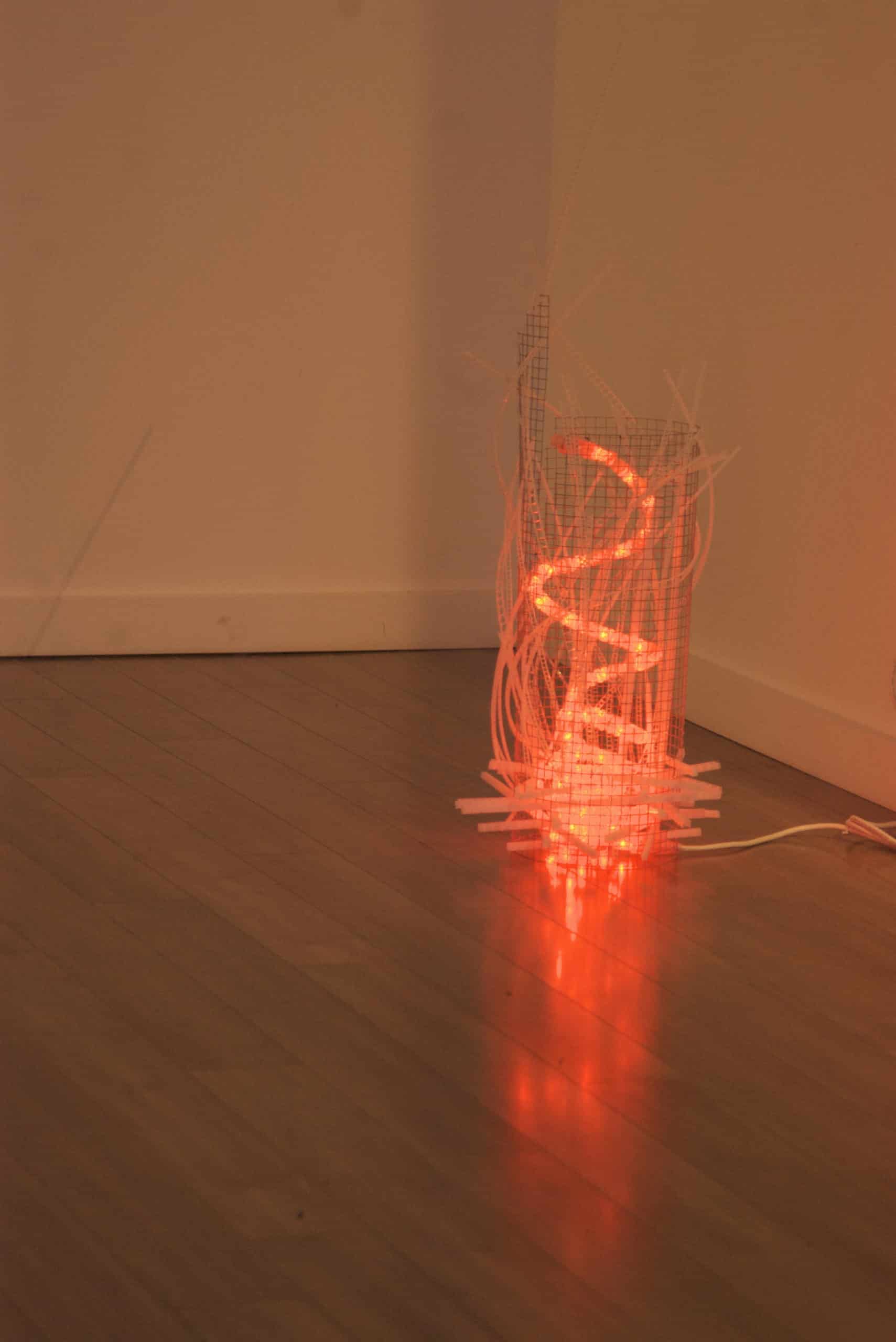 Campfire, freestanding red-orange original handmade Lumencrafter LED light sculpture, a swirling fiery rope light contained in a wire mesh cylinder shot through with strips of laddered polycarbonate twinwall on floor at Arcadia Art Gallery, Toronto, 2014.