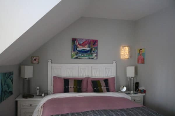 Box #1 original Lumencrafter wall sconce light sculpture mounted on bedroom wall surrounded by original paintings by Susette Godin.  Rectangular white battery-powered LED light sculpture, soft white fairy lights shine behind a diffusion plate framed by a sturdy steel grid with several thin twinwall strips poking through, up and down, back and forth.