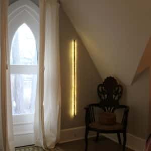 A one-inch wide strip of gold runs down the centre of a slender four-foot-long structure of polycarbonate struts and spars shot through with sparkling soft-white light.  Wall sculpture mounted in bedroom.    Original LED light art.