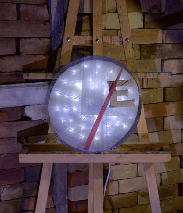 Runnin' On, original circular LED wall sconce light sculpture: A cool white LED rope light shines through a diffusion circle with large ceramic E and painted red pointer; polycarbonate strips poke through surrounding steel mesh cylinder. Sculpture rests comfortably on painter's easel.