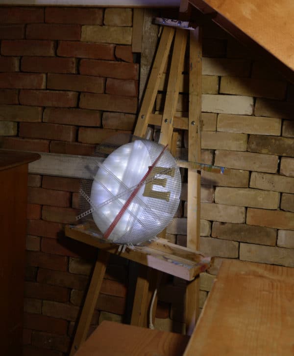 Runnin' On, original circular LED wall sconce light sculpture: A cool white LED rope light shines through a diffusion circle with large ceramic E and painted red pointer; polycarbonate strips poke through surrounding steel mesh cylinder. Sculpture rests comfortably on painter's easel under stairs.