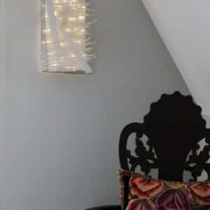 Box #1 original Lumencrafter wall sconce light sculpture mounted on bedroom wall by chair.  Rectangular white battery-powered LED light sculpture, soft white fairy lights shine behind a diffusion plate framed by a sturdy steel grid with several thin twinwall strips poking through, up and down, back and forth.