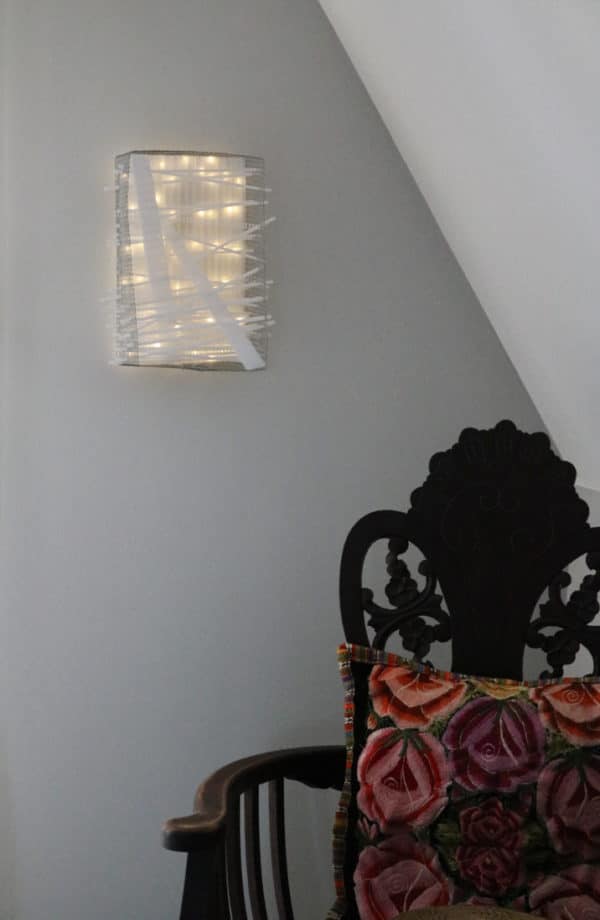 Box #1 original Lumencrafter wall sconce light sculpture mounted on bedroom wall by chair.  Rectangular white battery-powered LED light sculpture, soft white fairy lights shine behind a diffusion plate framed by a sturdy steel grid with several thin twinwall strips poking through, up and down, back and forth.