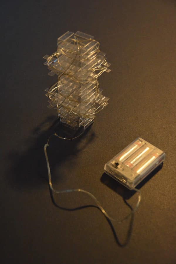 Clear inner core of  free-standing LED table light by Lumencrafter wrapped with LED light strip attached to battery pack.   Light yet sturdy, 6 inches high by four inch square base. Hand-painted plates of polycarbonate enclose dew drop LEDs wrapped around an inner polycarbonate core.  Indoor/outdoor. Suitable for evening al fresco dining or bedside mood lighting. Cordless; 3 AA batteries included.