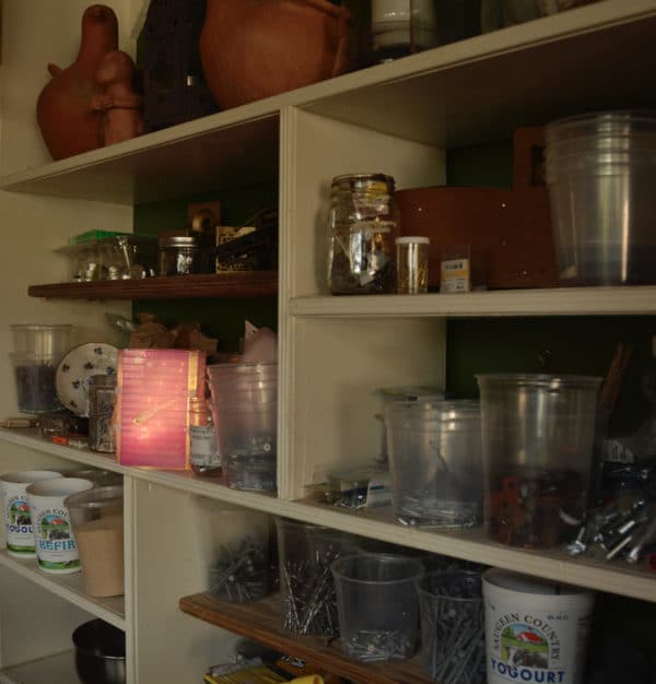 Free-standing LED table light by Lumencrafter, light pink with golden comet-splotch shines out from a shelf of bits and bobs.  Light yet sturdy, 6 inches high by four inch square base. Hand-painted plates of polycarbonate enclose dew drop LEDs wrapped around an inner polycarbonate core.  Indoor/outdoor. Suitable for evening al fresco dining or bedside mood lighting. Cordless; 3 AA batteries included.