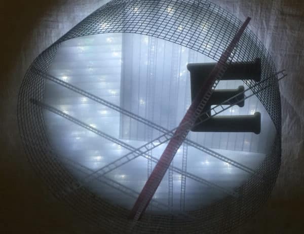 Runnin' On, original circular LED wall sconce light sculpture by Lumencrafter: A cool white LED rope light shines through a diffusion circle with large ceramic E and painted red pointer; polycarbonate strips poke through surrounding steel mesh cylinder. Sculpture shown at 2018 Grey County Autumn Leaves Studio Tour.