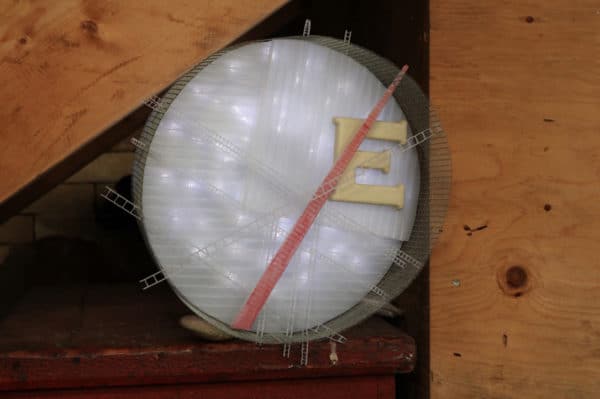 Runnin' On, original circular LED wall sconce light sculpture: A cool white LED rope light shines through a diffusion circle with large ceramic E and painted red pointer; polycarbonate strips poke through surrounding steel mesh cylinder. Sculpture rests on red wooden chest under stairs.