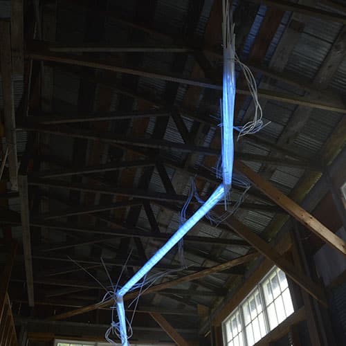A Roar, A Boar y Alice, a 22-foot-long original LED light sculpture by Lumencrafter, glows sky-blue as it hangs from the rafters of a driveshed. LED lights run through three acrylic tubes joined with heavy gauge wire, wrapped with strips of steel mesh and polycarbonate.  stunning; completely original.  cycles through a changing palette of glowing colour.